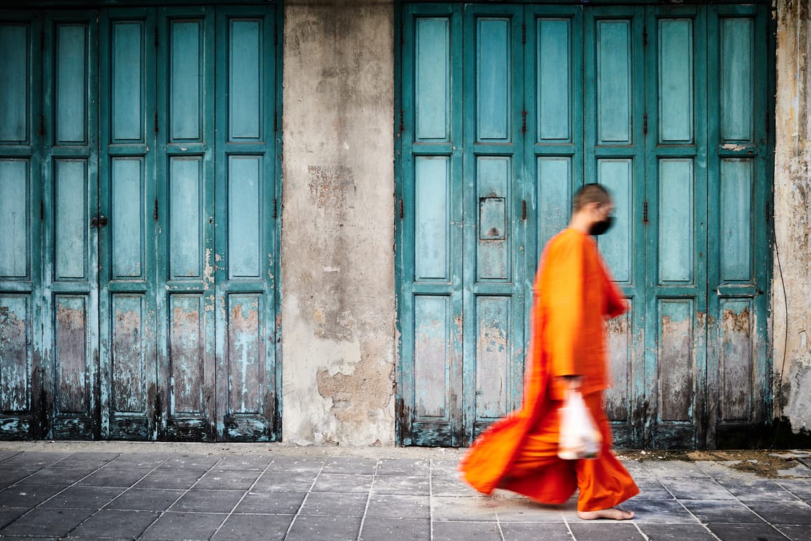 Un moine bouddhiste en robe orange vif passant devant les vieux stores verts en bois d’un magasin fermé.