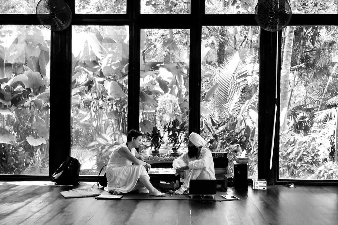 Deux personnes en tenue blanche discutent devant de grandes fenêtres avec vue sur un jardin tropical. Au centre, deux petites sculptures.